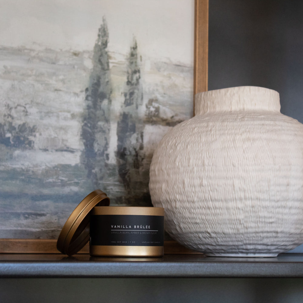 White textured vase on a shelf with a painting in the background with a gold candle tin