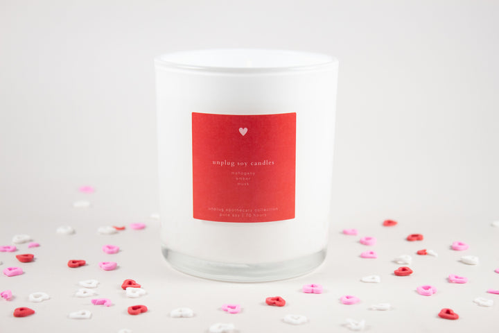 White candle with a red label on a light gray background with heart-shaped confetti.