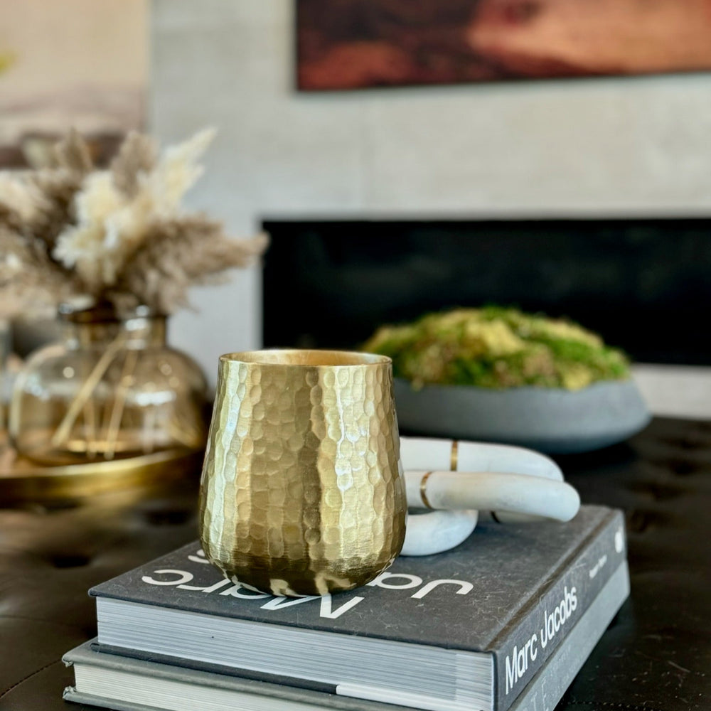 A gold hammered candle placed on top of a book, with a white decor item to its left and a glass vase with dried flowers in the background.