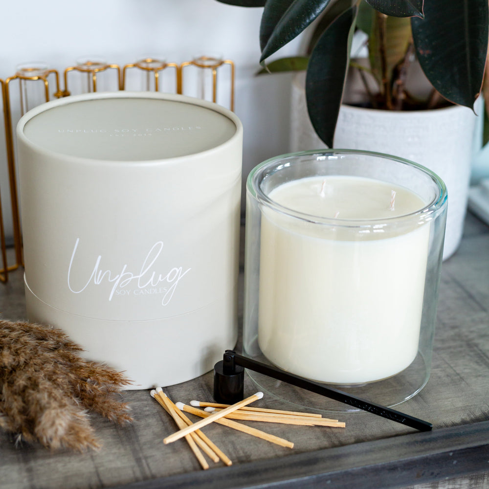 A large clear glass soy candle with a white label that reads 'Unplug', accompanied by a pair of black wicks and several wooden wicks on a table, with a decorative plant in the background.