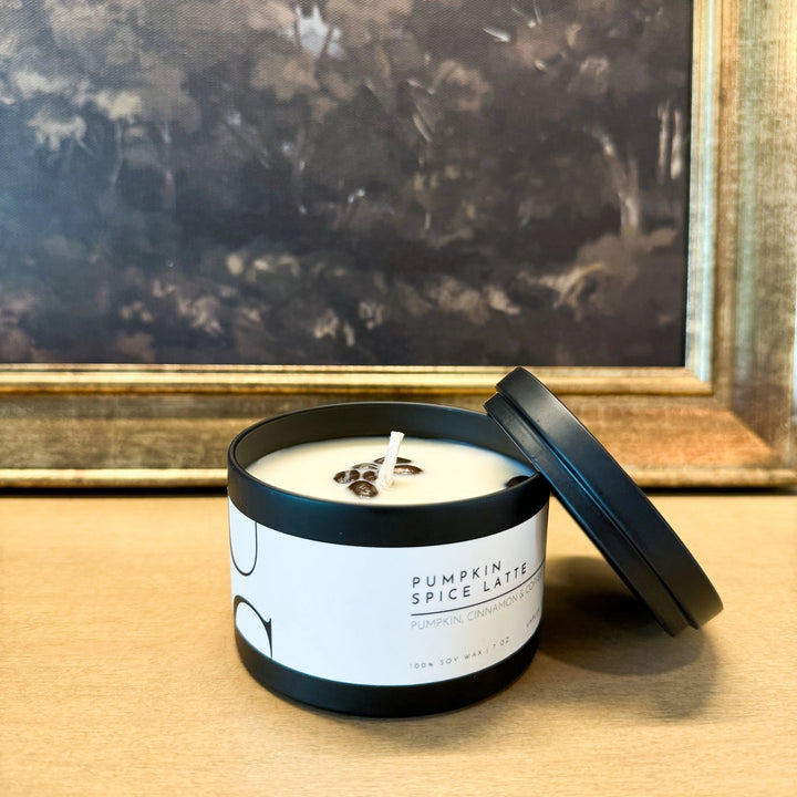 A matte black tin candle with the lid off, labeled 'Pumpkin Spice Latte', placed on a flat surface with a blurred picture frame in the background.